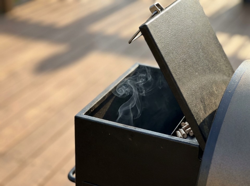 Close-up of a pellet grill's open hopper lid with a thin wisp of smoke curling out